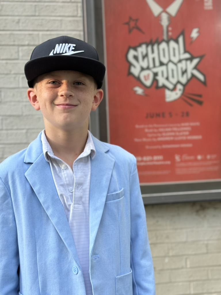 Holden in a black cap and light blue blazer in front of a School of Rock poster